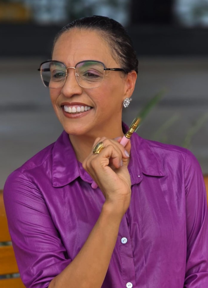 Teacher Dra. Izabel Borja sorrindo, de camisa roxa, segurando uma caneta